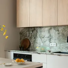 Jasna kuchnia z drewnianymi szafkami i kamiennym blatem. Wazon z żółtymi kwiatami stoi na wyspie. Co między szafkami w kuchni? Elegancki, kamienny backsplash i czarny kran.