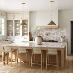 Jasna kuchnia w stylu modern classic z wyspą i czterema hokerami. Jasne meble, złote detale i duża ilość światła tworzą przytulną atmosferę.