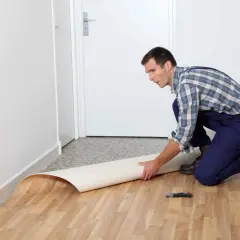 Mężczyzna w kombinezonie układa rolkę linoleum na podłodze. Pokazano, jak położyć linoleum na płytki.