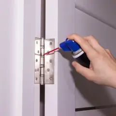 Dłoń z puszką preparatu smaruje zawias drzwi, pokazując, jak naoliwić drzwi, by przestały skrzypieć.