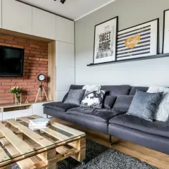 Nowoczesny salon z cegłą i drewnem. Szara kanapa, stolik z palet i grafiki na ścianie tworzą przytulną atmosferę.