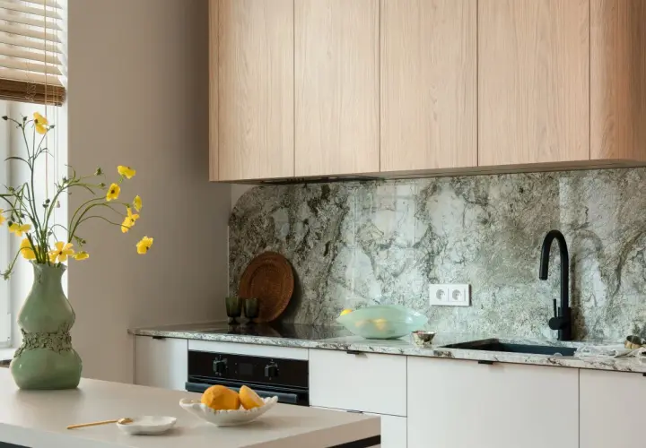 Jasna kuchnia z drewnianymi szafkami i kamiennym blatem. Wazon z żółtymi kwiatami stoi na wyspie. Co między szafkami w kuchni? Elegancki, kamienny backsplash i czarny kran.