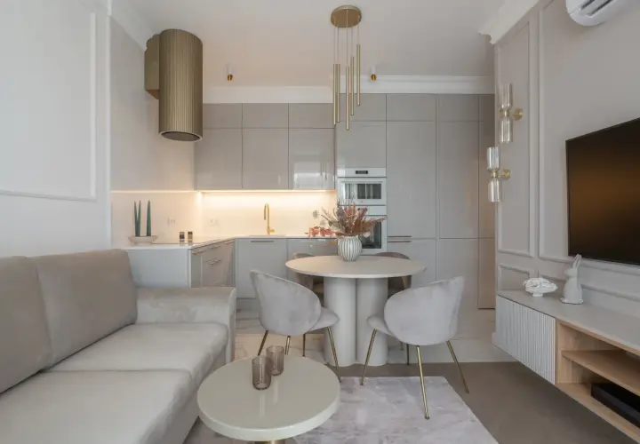 Elegancka, mała kuchnia z salonem. Jasna kanapa, okrągły stół z krzesłami i nowoczesna zabudowa kuchenna tworzą przytulną przestrzeń.