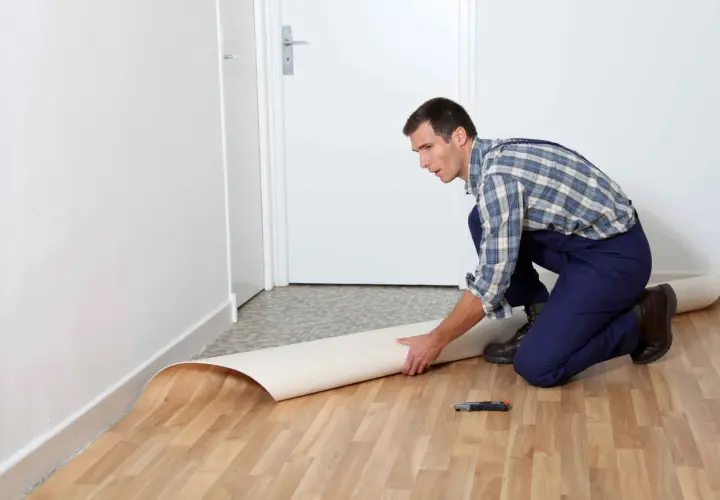 Mężczyzna w kombinezonie układa rolkę linoleum na podłodze. Pokazano, jak położyć linoleum na płytki.