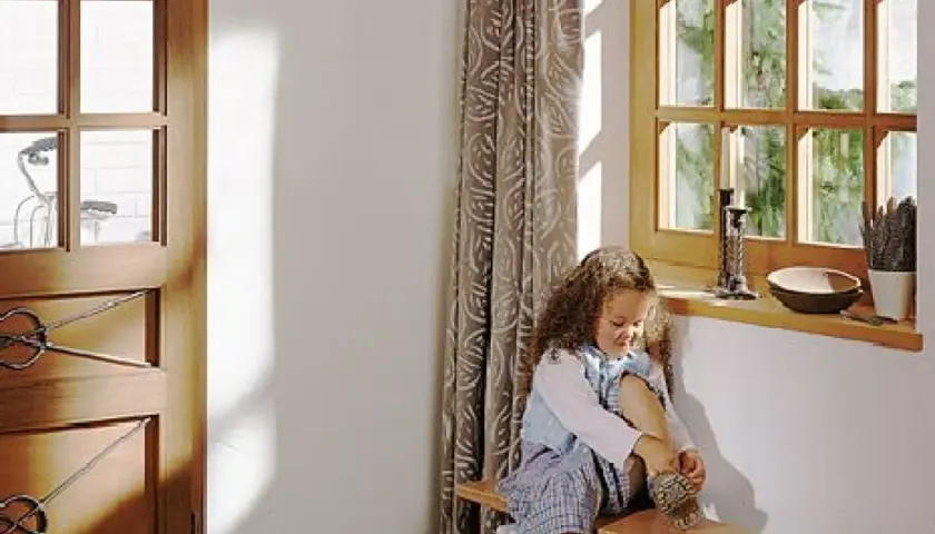 Dziewczynka w sieni zakłada buty. Obok okno z widokiem na ogród i drzwi z ozdobnymi okuciami.