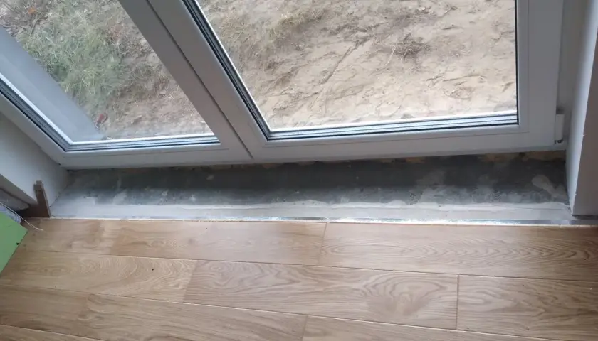 Drewniane panele podłogowe przy drzwiach balkonowych, gotowe do wykończenia. Widoczny betonowy próg i fragment okna.