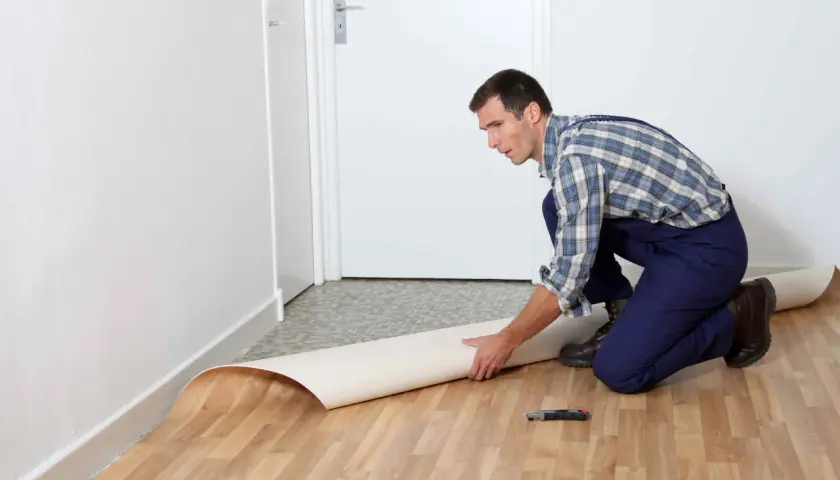 Mężczyzna w kombinezonie układa rolkę linoleum na podłodze. Pokazano, jak położyć linoleum na płytki.