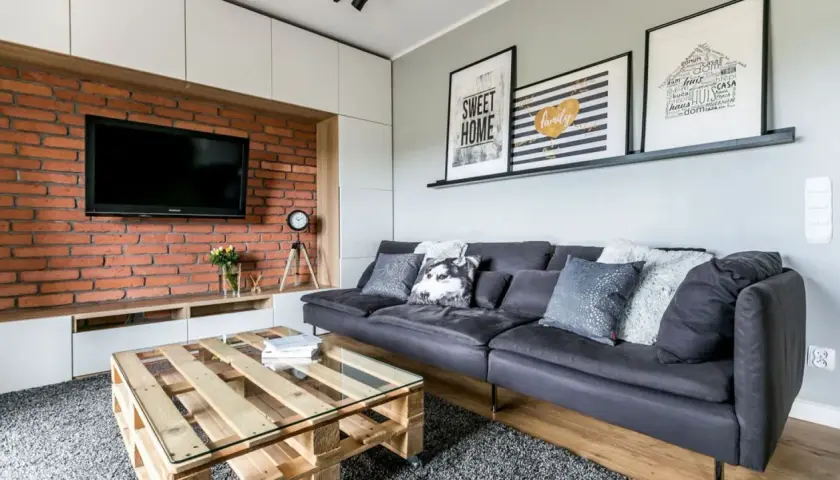 Nowoczesny salon z cegłą i drewnem. Szara kanapa, stolik z palet i grafiki na ścianie tworzą przytulną atmosferę.