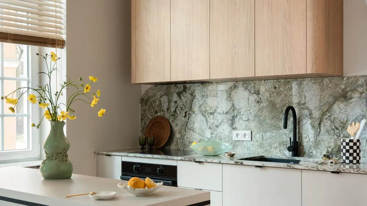 Jasna kuchnia z drewnianymi szafkami i kamiennym blatem. Wazon z żółtymi kwiatami stoi na wyspie. Co między szafkami w kuchni? Elegancki, kamienny backsplash i czarny kran.