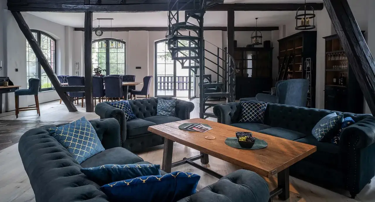 Wnętrze w stylu industrialnym z ciemnymi sofami, drewnianym stołem i krętymi schodami.