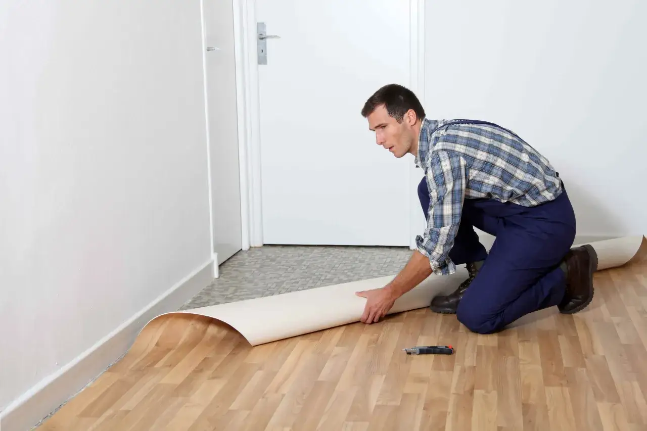 Mężczyzna w kombinezonie układa rolkę linoleum na podłodze. Pokazuje, jak położyć linoleum na płytki.